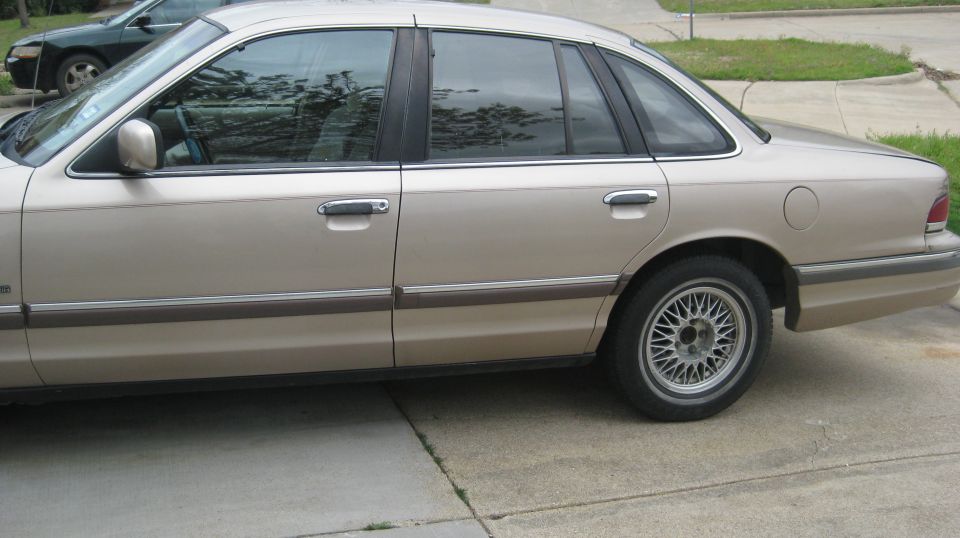 Side view of a tan sedan parked on a driveway. Another dark car is visible in the background