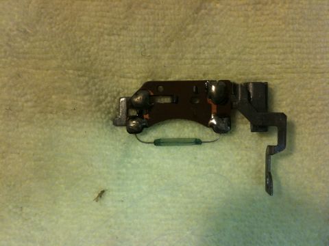 Suzuki Samurai - Small stamped-steel latch or lock mechanism laid on a yellow shop towel, showing four riveted pivot posts, a slotted plate, a green coil re…