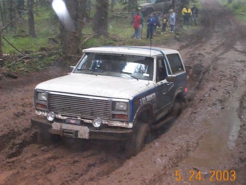 1983 Ford Bronco - A Ford Bronco navigates a muddy off-road trail with onlookers in the background.