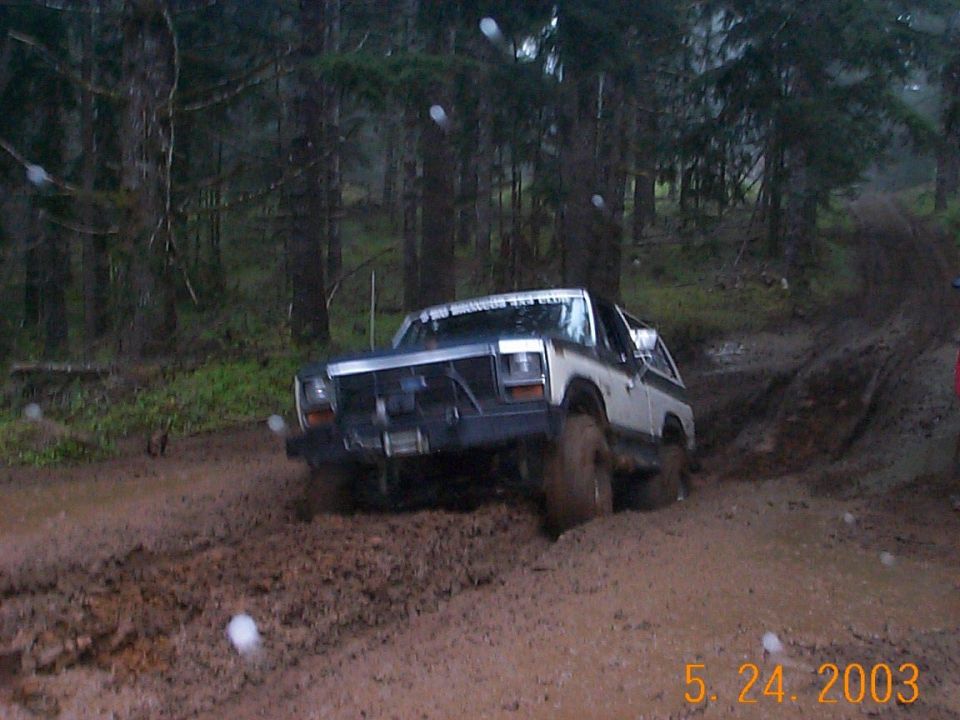 Silver Ford Bronco climbing a muddy forest trail, mud-splattered with brush guard and winch.