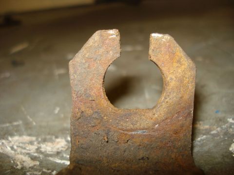 1979 Ford Bronco - Extreme close-up of a rusty stamped-steel bracket standing on a workbench, its top cut into a two-pronged fork with a round keyhole slot fo…
