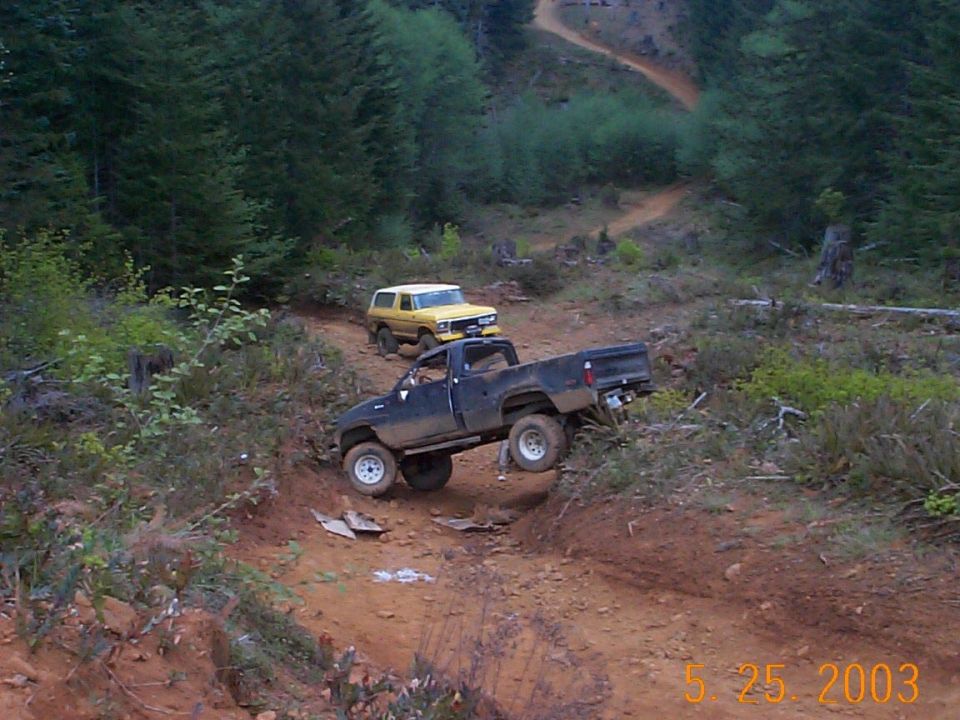 Two lifted 4x4 trucks on a muddy off-road trail cut through evergreen forest