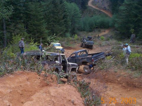 1983 Ford Bronco - Multiple off-road vehicles traverse a challenging dirt path surrounded by dense forest.