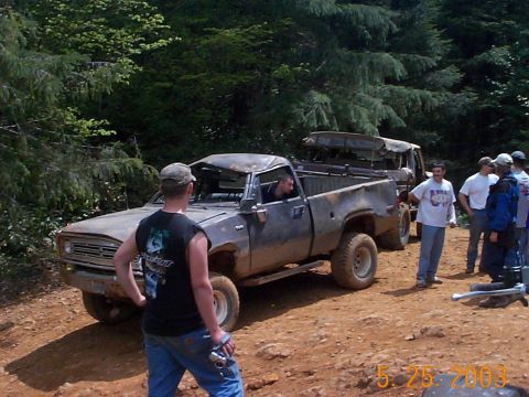 1983 Ford Bronco - Several people gather around a heavily damaged pickup truck on a wooded dirt path.