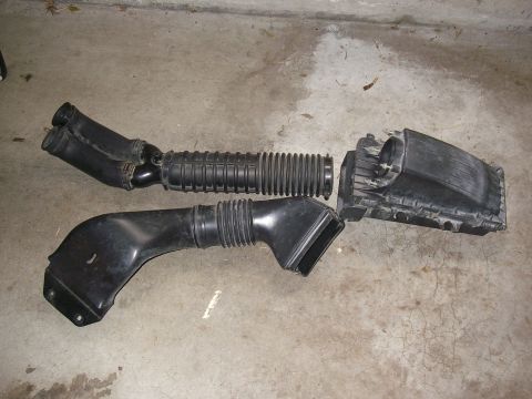 1990 Ford Bronco - Removed engine air intake parts including ribbed ducting and the air filter box resting on a concrete floor.