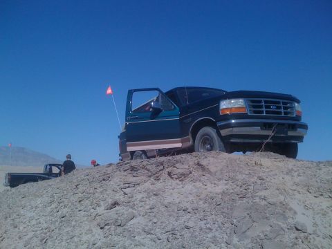 1995 Ford Bronco - A dark green Ford Bronco is parked on a sandy hill with its door open, under a clear blue sky.