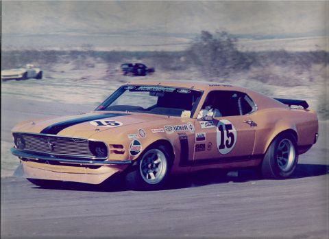1973 Ford Mustang - A vintage race car with sponsor decals speeds around a track, with other vehicles visible in the distance.