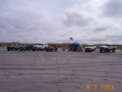 1983 Ford Bronco - Multiple SUVs and trucks are parked on a sandy beach under a cloudy sky.