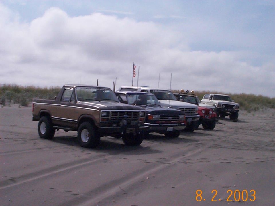 Five 4x4s lined up on beach sand, led by a tan 1980s Ford Bronco with top removed
