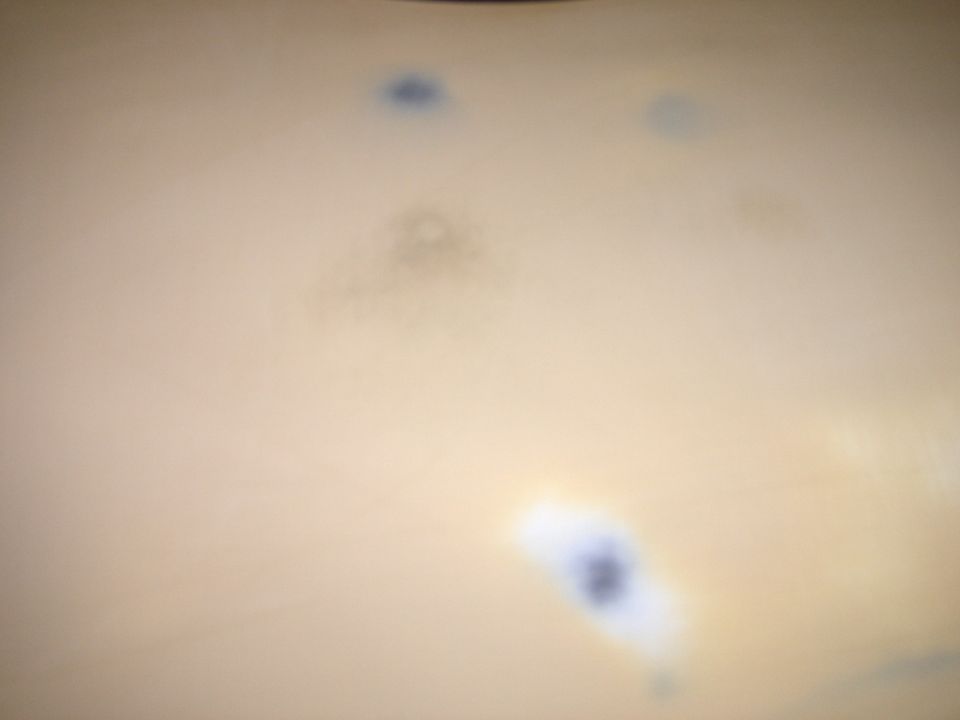 close-up of light-colored surface with blue and brown spots, likely paint flaws. Supermotors.net logo visible