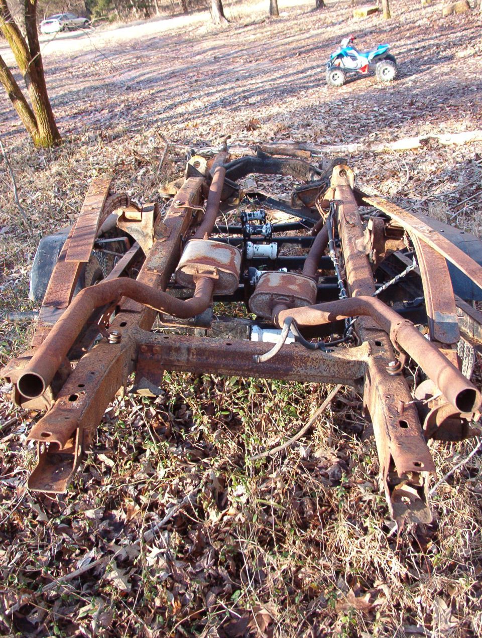 Rusty bare vehicle frame with exhaust and mufflers lying on leaf-covered ground, toy ATV in background