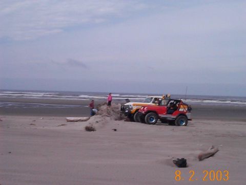 1983 Ford Bronco - Two off-road vehicles are parked on a beach with people nearby and the ocean in the background.