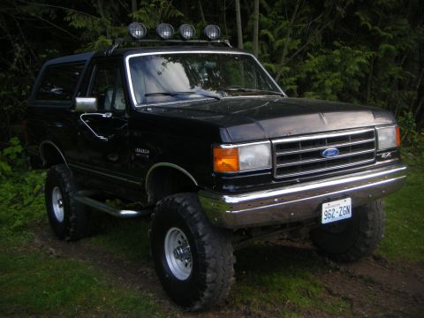 Black Ford Bronco lifted with off-road tires and roof lights, parked on a dirt path with trees in the backgro…