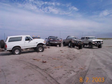 1983 Ford Bronco - Multiple off-road vehicles gathered on a sandy beach with a cloudy sky overhead.