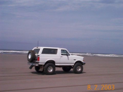 1983 Ford Bronco - A white SUV drives along a sandy beach with the ocean in the background.