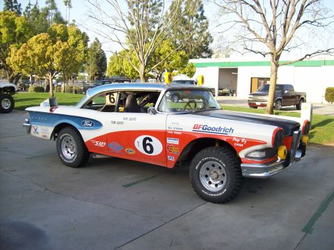 1990 Ford Bronco - Front three-quarter view of a vintage Edsel race car in orange, white and black livery on off-road tires parked among trees.