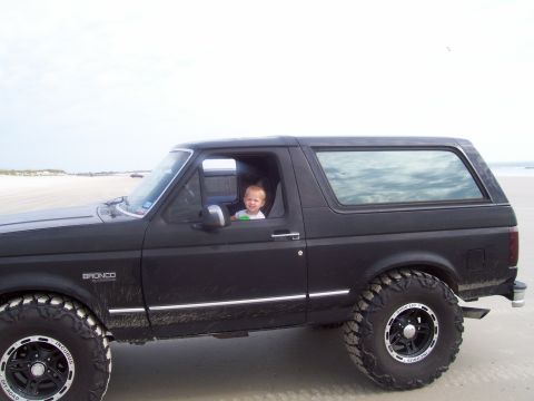1994 Ford Bronco - Child sits in a black Ford Bronco parked on a sandy beach with the ocean in the background.