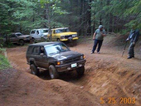 1983 Ford Bronco - Several off-road vehicles navigate a wooded trail, with a black SUV in the foreground.