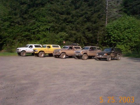 1983 Ford Bronco - A group of five SUVs, including a yellow Ford Bronco, are parked together in a gravel lot with a wooded area behind them.