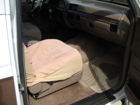 1993 Ford Bronco — Interior shot of a truck's passenger side, showing the seat with a cover, dashboard, and floor mat. Text on t…