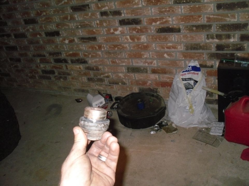 Hand holding a metal automotive part, possibly a bearing or bushing. Other items like a cast iron pot and a bag of absorbent are in the background