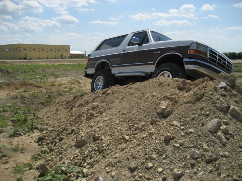 1993 Ford Bronco - This lifted 1993 Ford Bronco is shown off-road, navigating a dirt pile under a partly cloudy sky.
