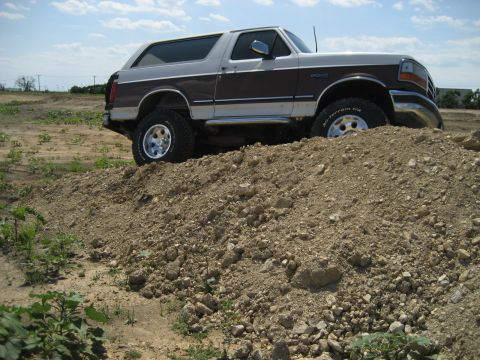1993 Ford Bronco - A lifted Ford Bronco navigates a dirt obstacle under a partly cloudy sky.