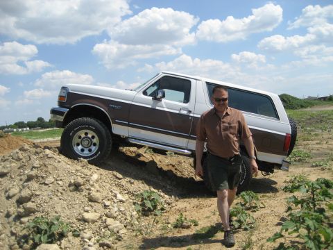 1993 Ford Bronco - A man walks past a white and brown 1993 Ford Bronco parked on a dirt incline under a cloudy sky.