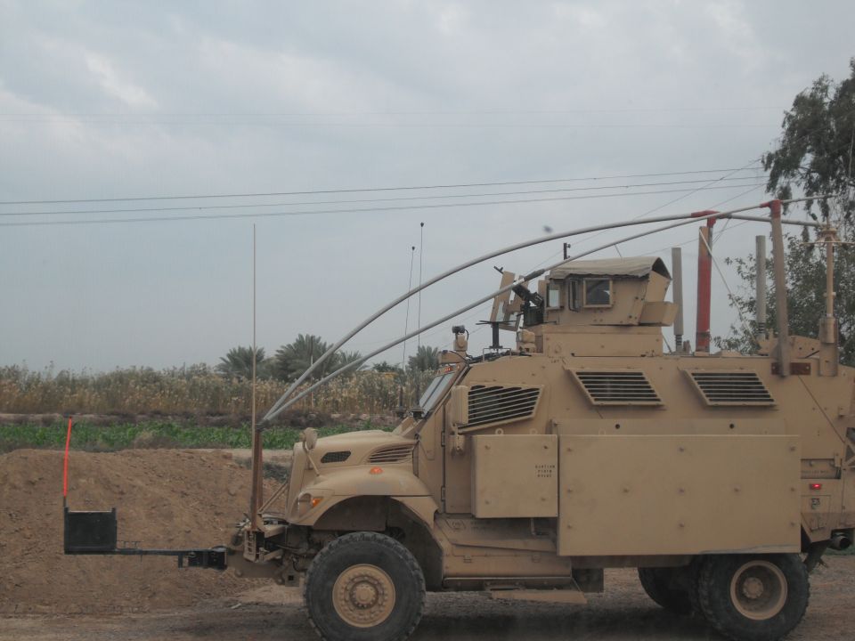 Tan military truck with a mounted weapon on top, parked in a dusty area with vegetation in the background