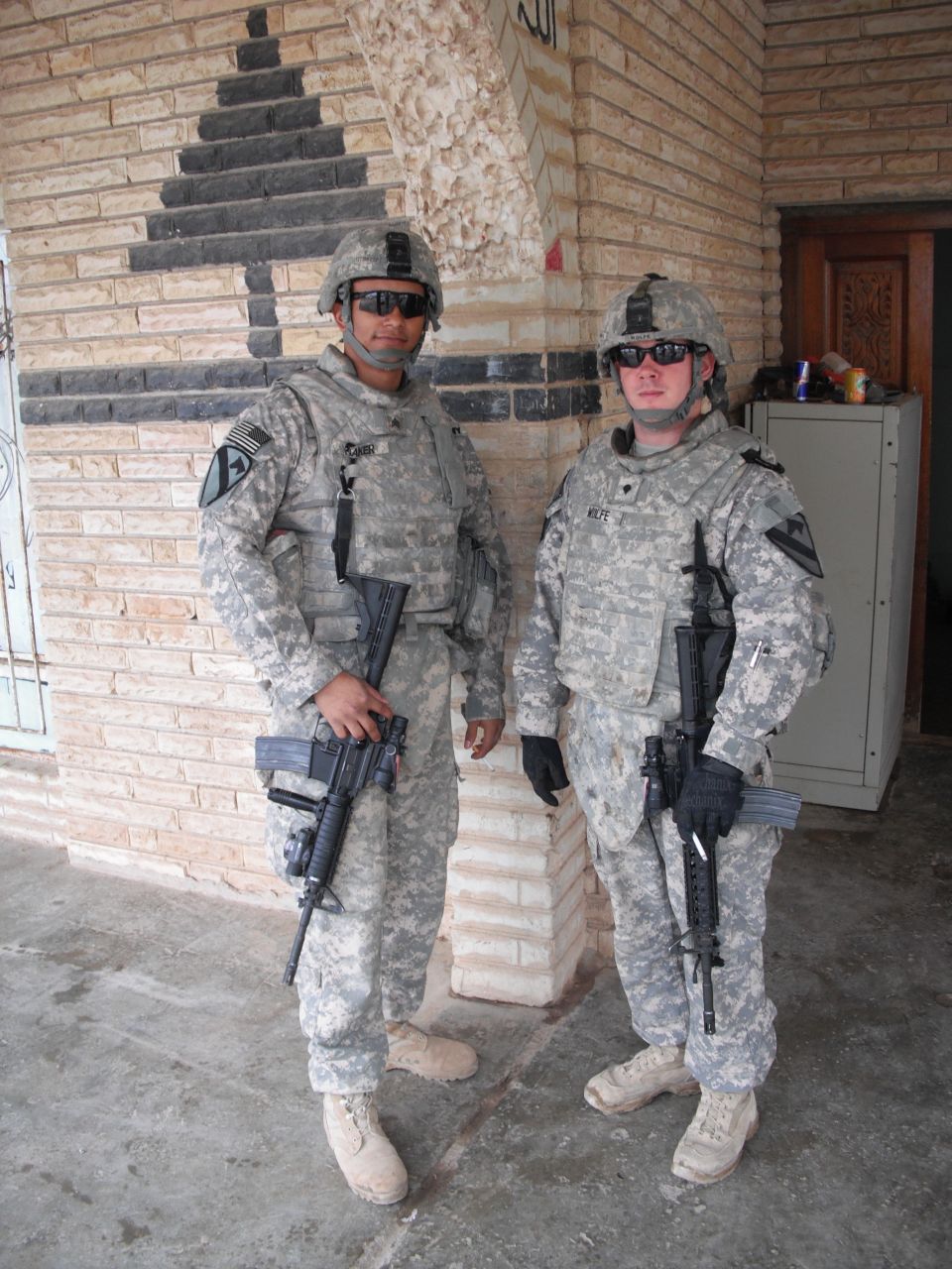Two soldiers in camouflage uniforms stand with rifles in front of a brick wall