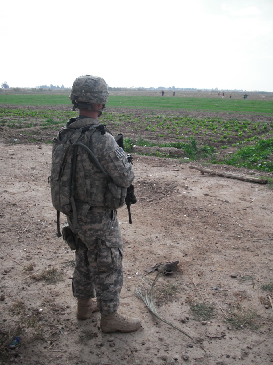 soldier standing in a dirt area looking out at a field with other soldiers in the distance
