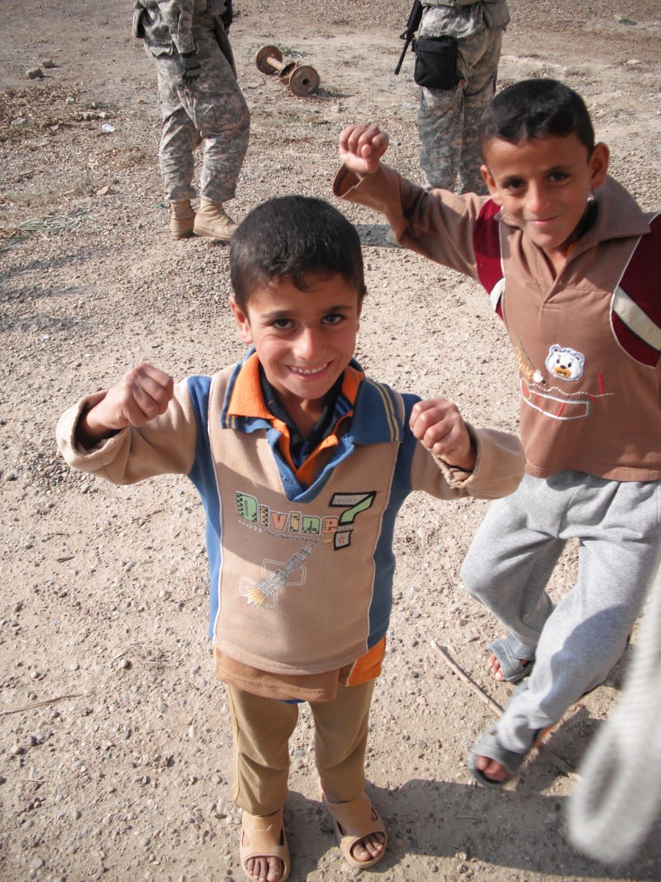 Two young boys in casual clothes pose with clenched fists in a dusty outdoor area. Soldiers are visible in the background