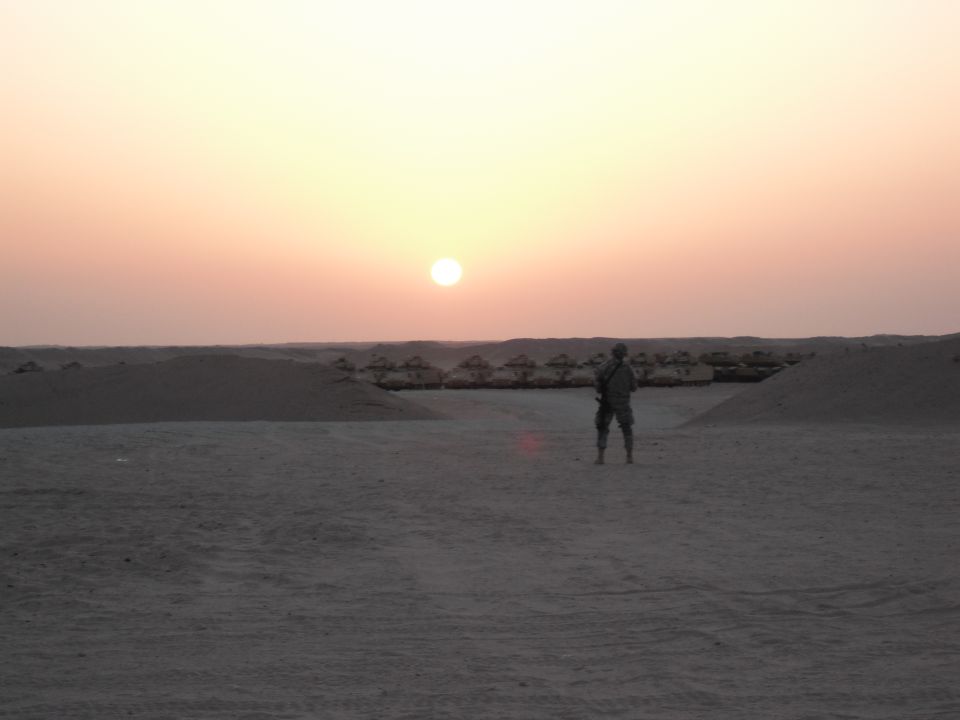 Soldier standing in desert with row of military vehicles behind him at sunset