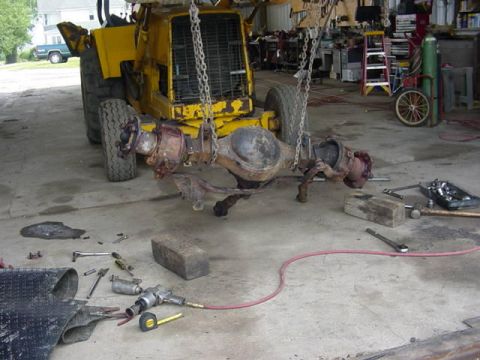 1992 Ford Bronco - A heavy planetary-hub axle assembly hangs from chains on a yellow forklift inside a shop, with both hub ends and the round center pumpkin e…