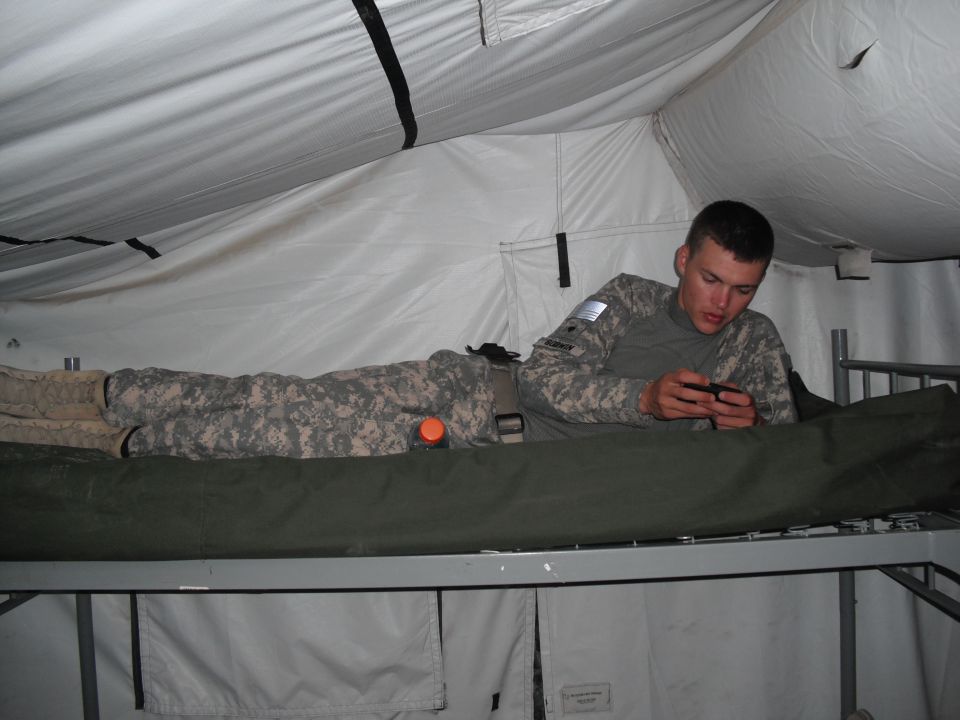 Soldier in camouflage uniform lying on a cot inside a tent, looking at his phone. Another person in uniform is lying down next to him