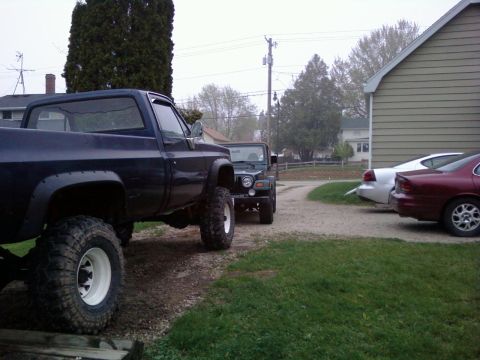 1988 Ford F-150 - A lifted pickup truck and a Jeep are parked on a gravel driveway next to a building and two sedans.