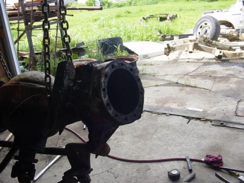 1992 Ford Bronco - A heavy cast-iron axle housing hangs from a chain hoist in a garage doorway during disassembly, its round differential carrier flange strip…