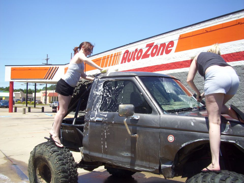 Two women washing a lifted, old-school pickup truck in front of an AutoZone