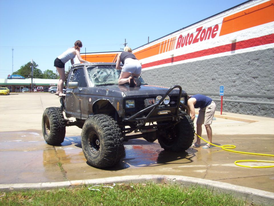 Dark-colored, lifted truck with large tires being washed by three people in front of an AutoZone