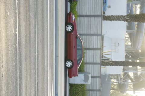 1992 Ford Taurus - A side view of a red 1992 Ford Taurus sedan is captured against a backdrop of a concrete wall and palm trees.