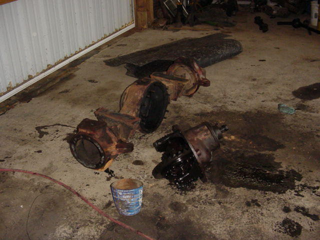 Disassembled Ford Bronco axles on a concrete floor with a bucket and oil stains