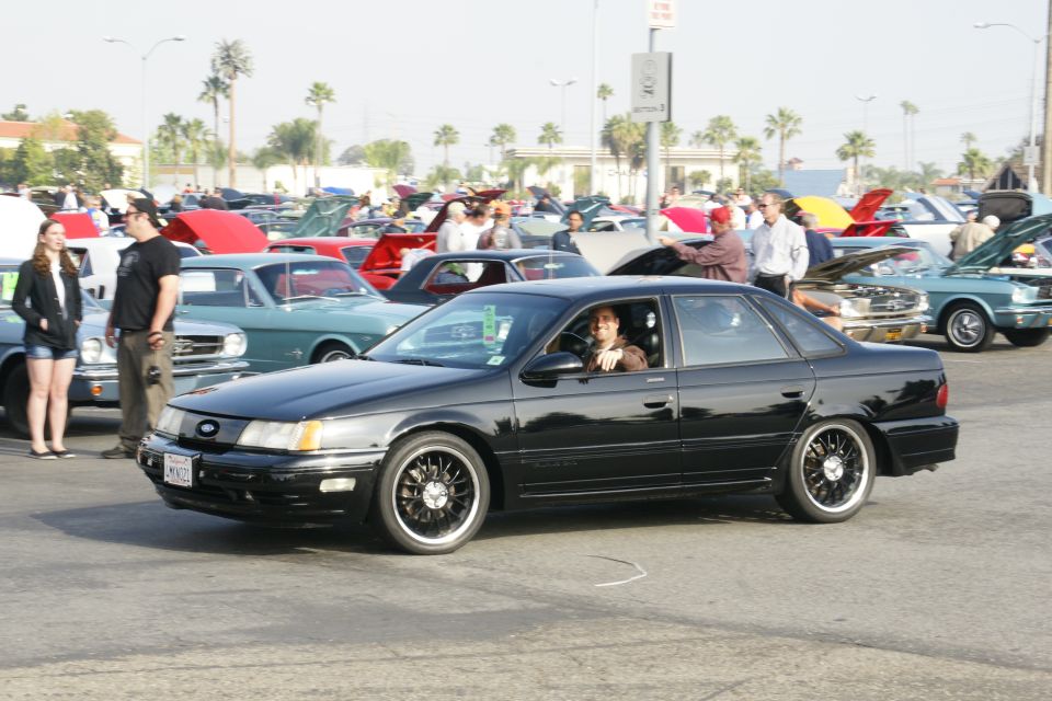 Black 1992 Ford Taurus sedan with custom wheels, driver smiling. Other cars in background