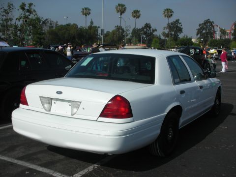 2004 Ford Crown Victoria - A white sedan is parked in a lot with other vehicles and people in the background.