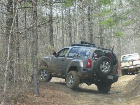 1993 Ford Bronco - Two off-road vehicles navigate a wooded trail, one gray SUV and a beige pickup truck.