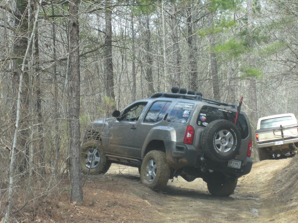 Gray SUV with spare tire on back and beige pickup truck on dirt trail in woods