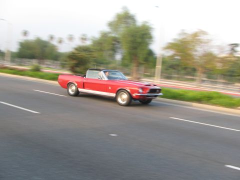 1996 Ford C-350 (Centurion Conversions) - A red 1967 Ford Mustang convertible cruises down a highway, its classic lines blurred by motion.
