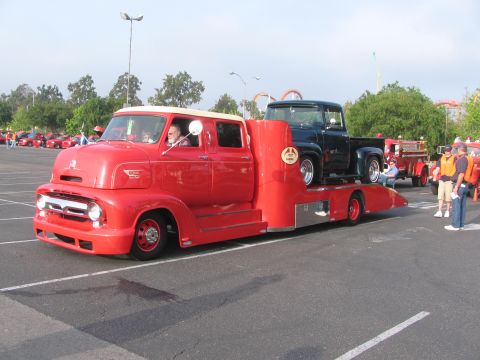1996 Ford C-350 (Centurion Conversions) - A red tow truck transports a dark green 1956 Ford F-100 pickup truck, with other vehicles visible in the background.