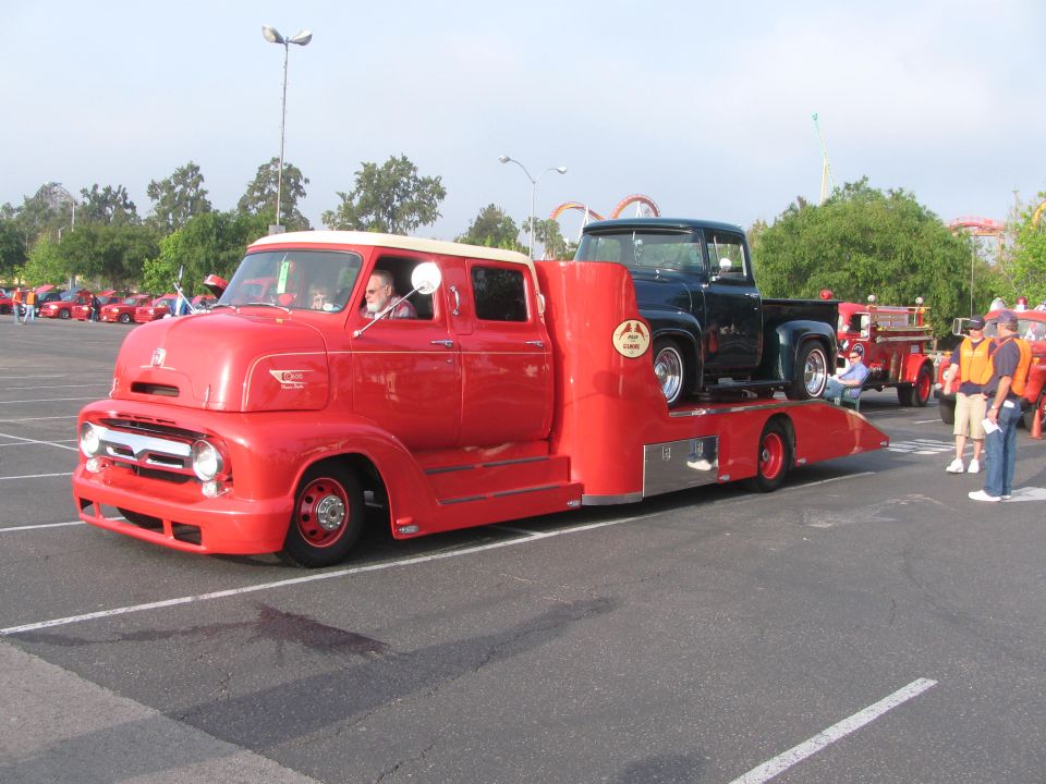 Red tow truck carrying a dark green 1956 Ford F-100 pickup truck