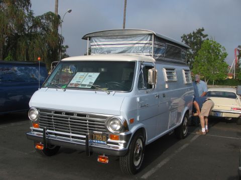 1996 Ford C-350 (Centurion Conversions) - A light blue Ford Econoline camper van with a pop-top roof is parked in a lot, with two people near the rear.