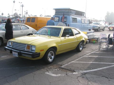 1996 Ford C-350 (Centurion Conversions) - A yellow 1977 Ford Pinto coupe is parked in a lot with other vehicles, including a blue camper van and a yellow van.